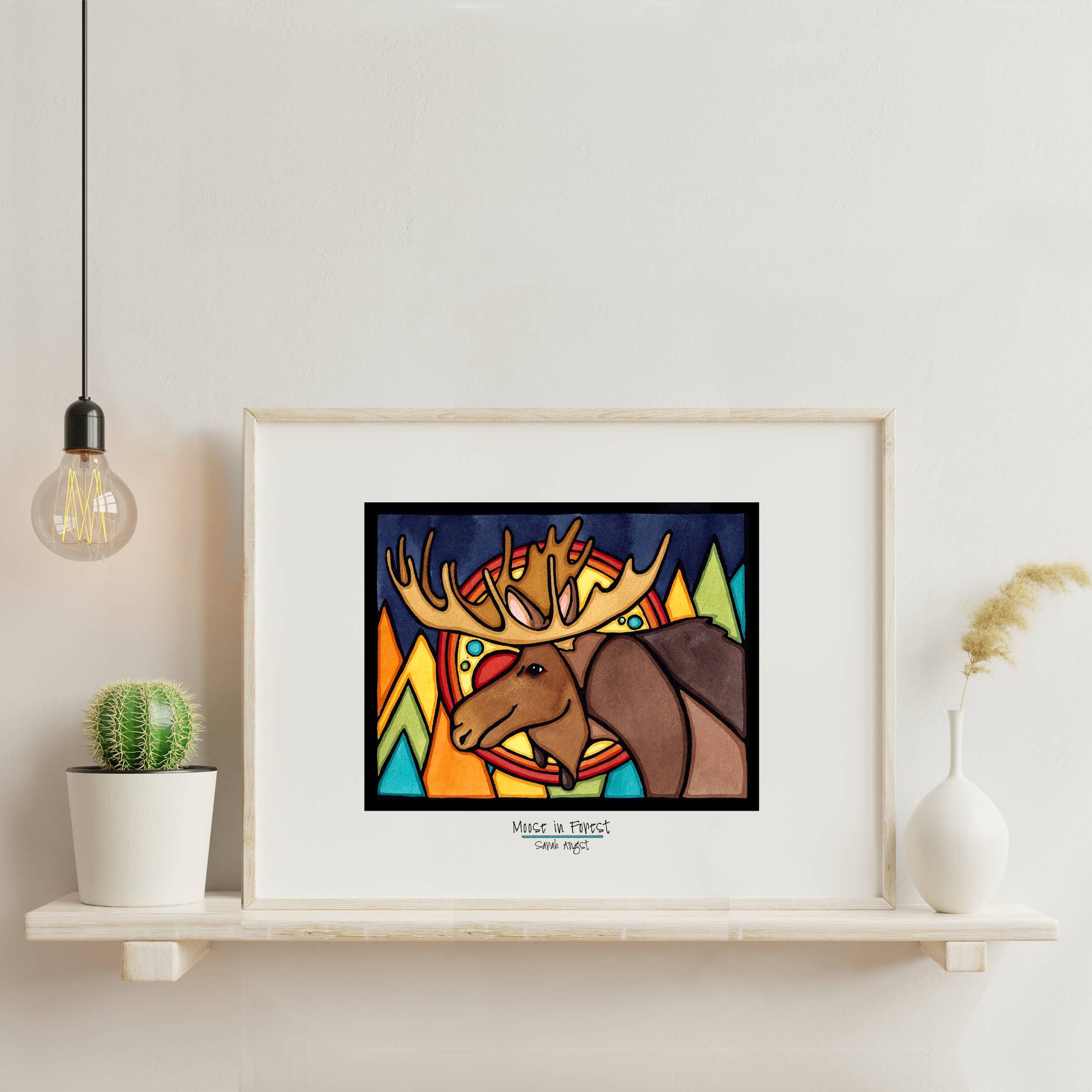 The Artwork: Moose in Forest: 11"x14" by Sarah Angst sits on a shelf alongside a small cactus, bulb light, and dried plant. This framed illustration is a vibrant giclee art print that beautifully captures the essence of nature with stunning detail.