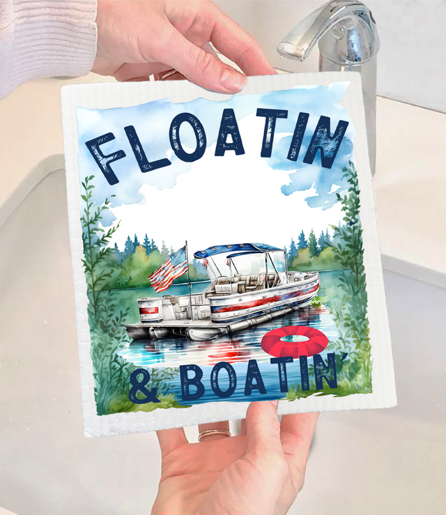 A person holds the Dish Cloth: Floating and Boating on the Lake Swedish Dishcloth, which features a pontoon boat on a lake, an American flag, and bold blue text reading FLOATIN & BOATIN.