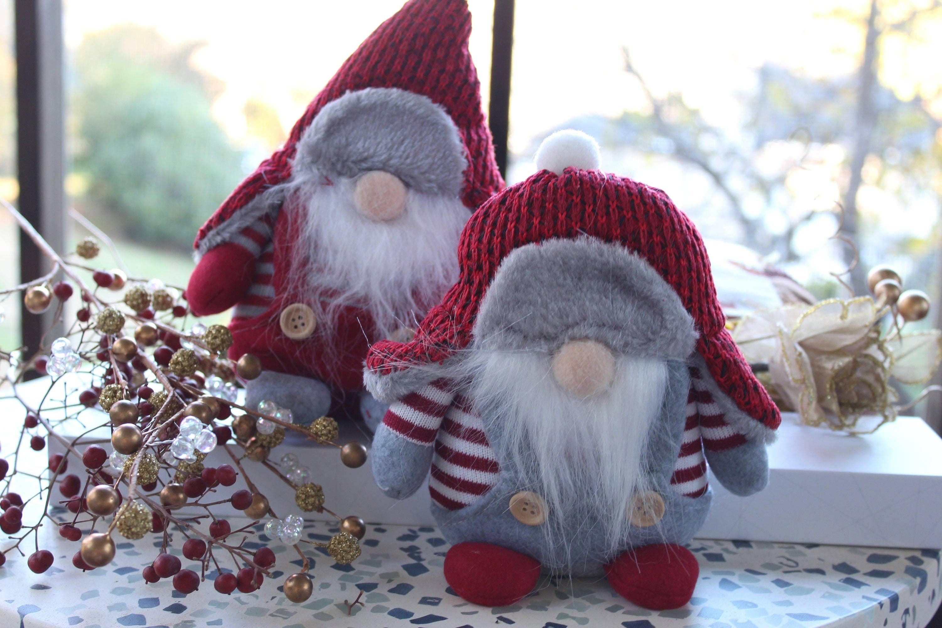 Two Aldrich or Bernard Christmas Gnomes, donned in red hats and striped shirts inspired by Scandinavian folklore, are seated on a patterned surface. A festive branch is positioned close by.