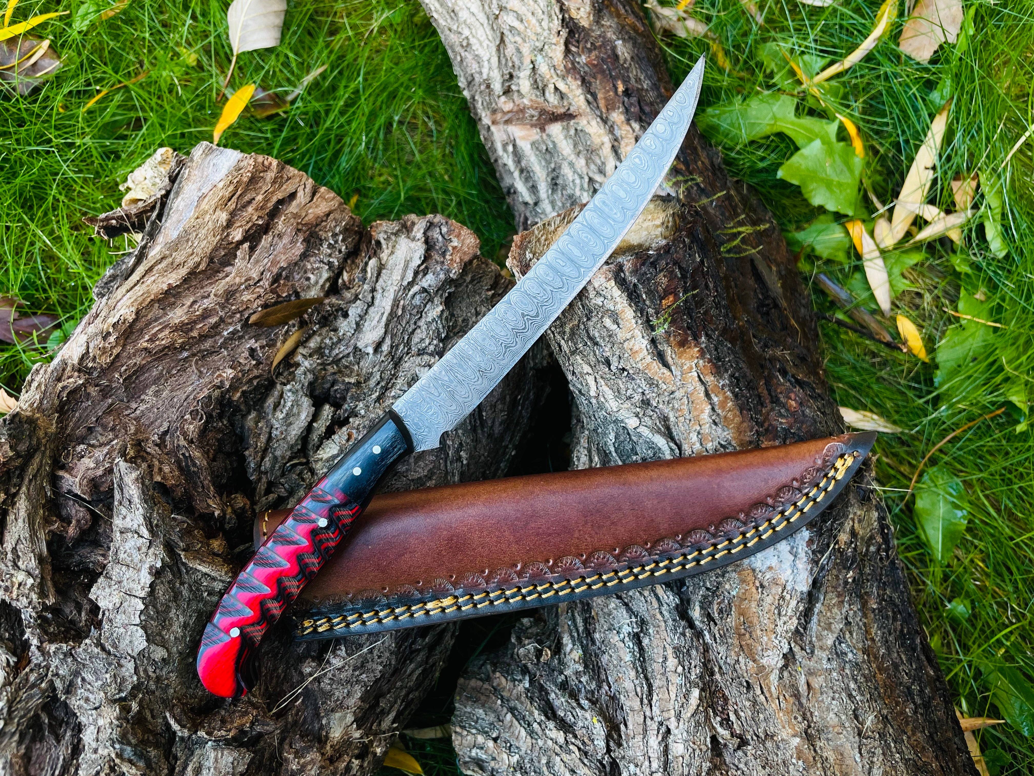 The Knife: Damascus Steel Custom Flay Knife with Craved Grips features a patterned steel blade and a red-black handle, displayed on wood beside its brown leather sheath, with grass and leaves in the background.