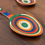 Birch serving spoon with green, orange, blue, red, and yellow rings on handle lying on a brown wooden surface; another birch spoon is partially visible in the background.