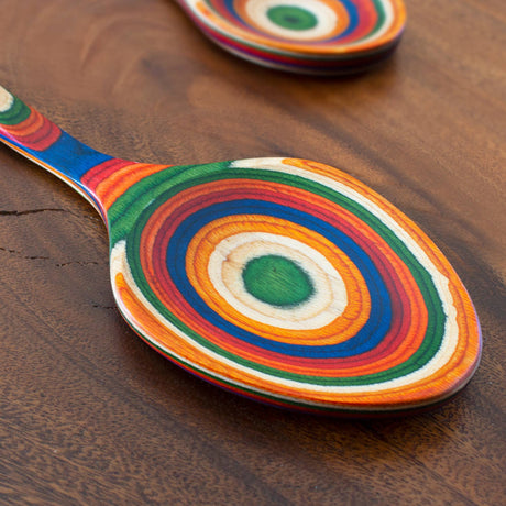 Birch serving spoon with green, orange, blue, red, and yellow rings on handle lying on a brown wooden surface; another birch spoon is partially visible in the background.