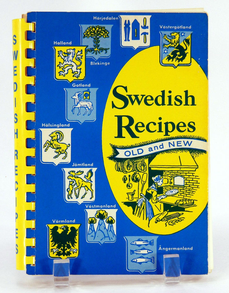 The 1955 edition of the book "Swedish Recipes Old & New" showcases its blue and yellow cover adorned with various heraldic symbols and a kitchen illustration, curated by the American Daughters of Sweden.