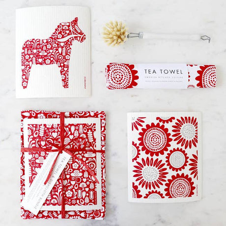 A collection of red and white kitchen items on a marble surface, featuring an environmentally friendly tea towel, a biodegradable brush, a wrapped package, and the Dish Cloth: Dala Horse Red card with a horse motif.