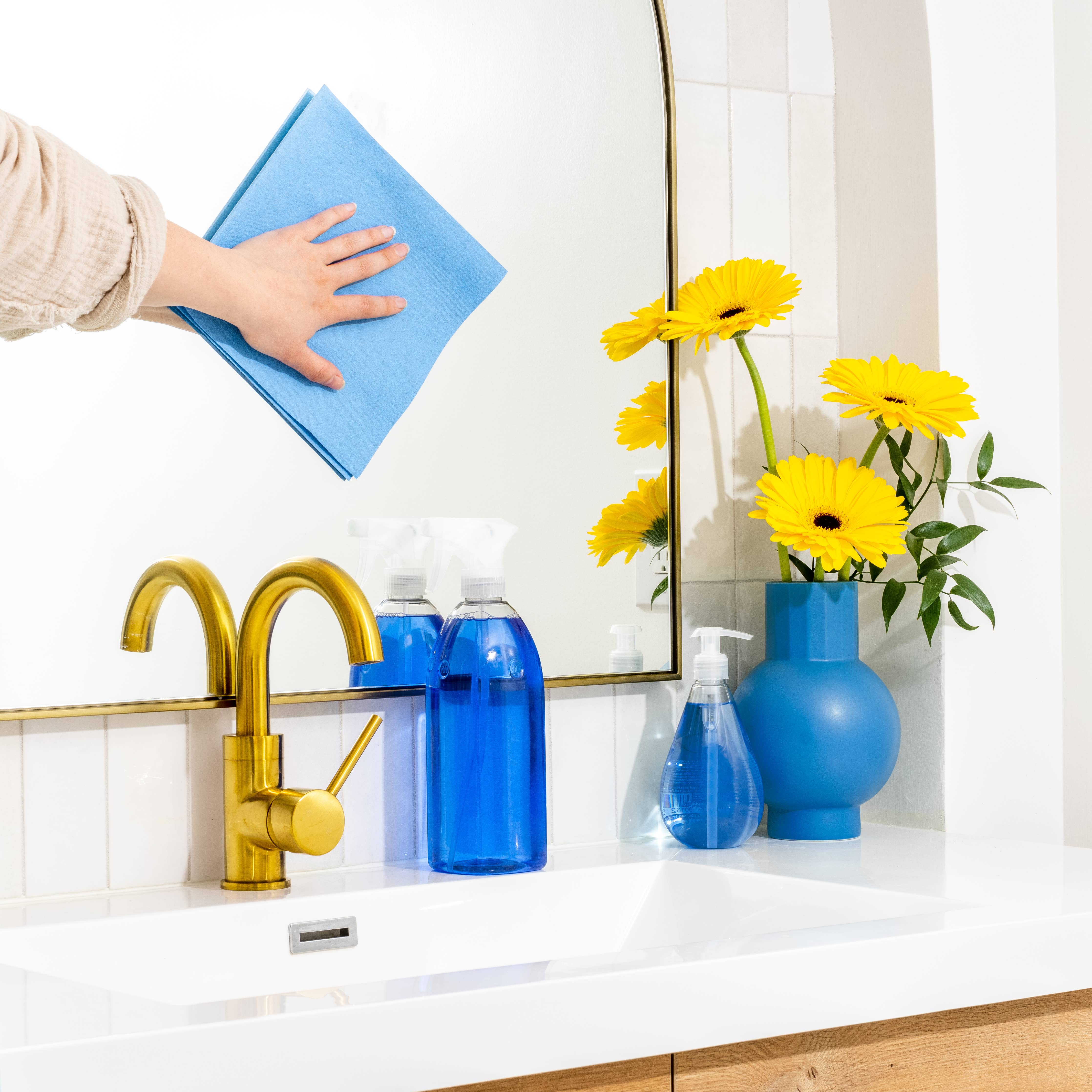 A hand uses the Streak Free Cloth Euroscrubby to clean a mirror above a sink, ensuring spotless results. Blue cleaning products and a blue vase with yellow flowers decorate the counter.