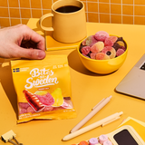 A hand holds a yellow bag of Swedish candy next to a yellow bowl of gummies, a mug, pens, and a laptop on a bright yellow desk.