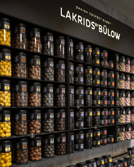 Wall display of large jars containing assorted colors and flavors of Lakrids by Bülow licorice, including varieties like salty licorice and white chocolate licorice.
