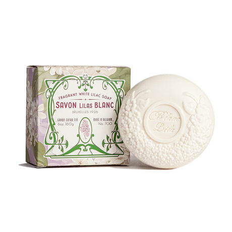 The round, white Blanc Lila Lilac Savon Bar Soap features floral embossing and is shown beside a decorative, plastic-free box with a green and lavender floral design labeled "Savon Lilas Blanc.