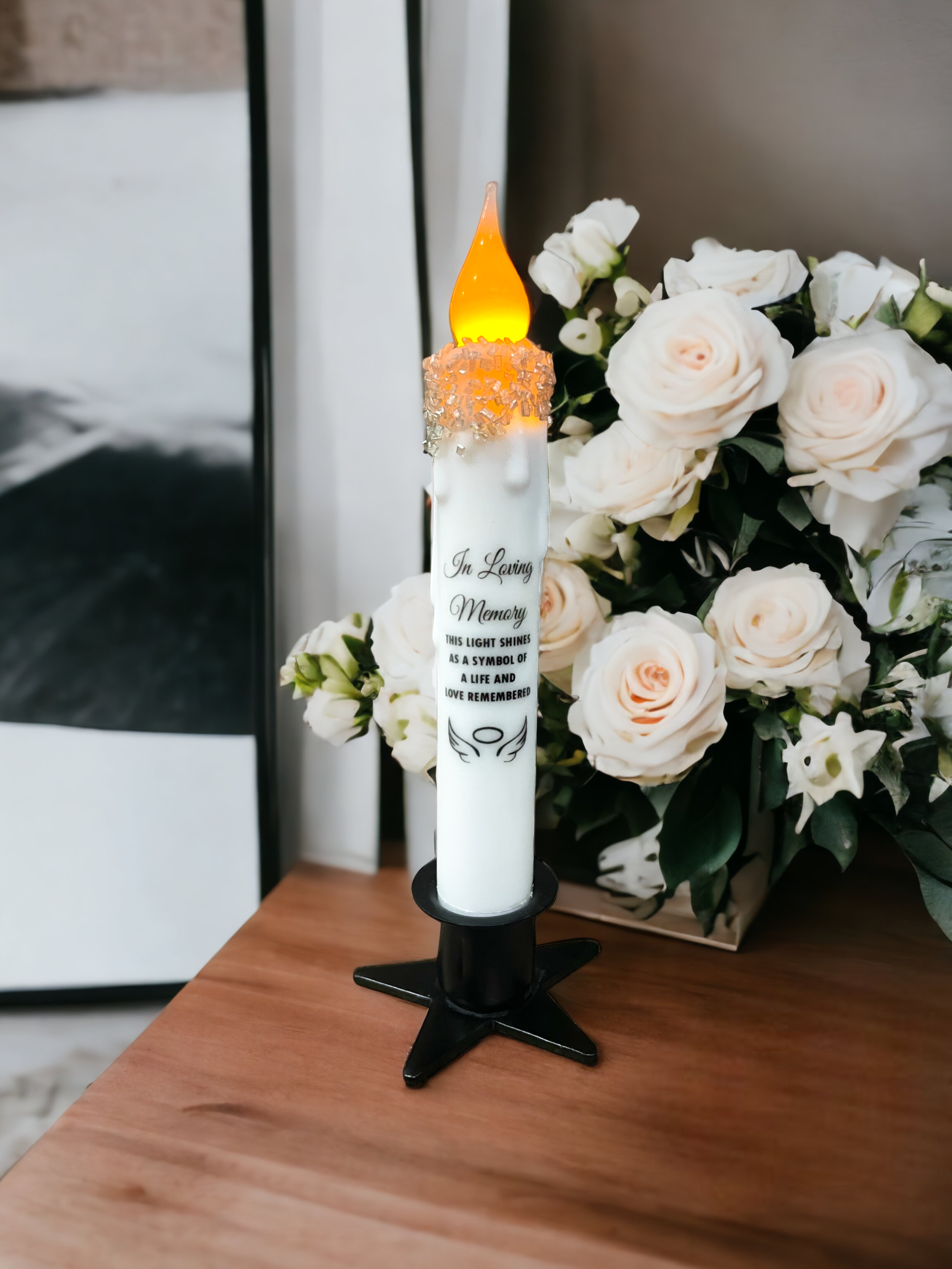 A Star Wrought Iron Taper Holder with a lit memorial candle and decorative beads stands in a candle display box beside a bouquet of white roses on a wooden surface.