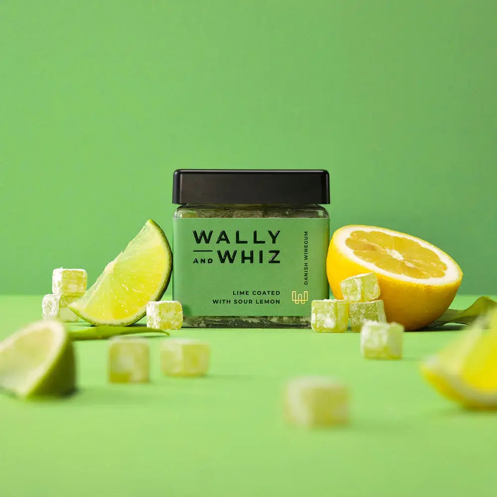 A 140g jar of Wally & Whiz Lime With Sour Lemon plant-based candy is displayed with lemon and lime wedges plus green cubes against a green background.