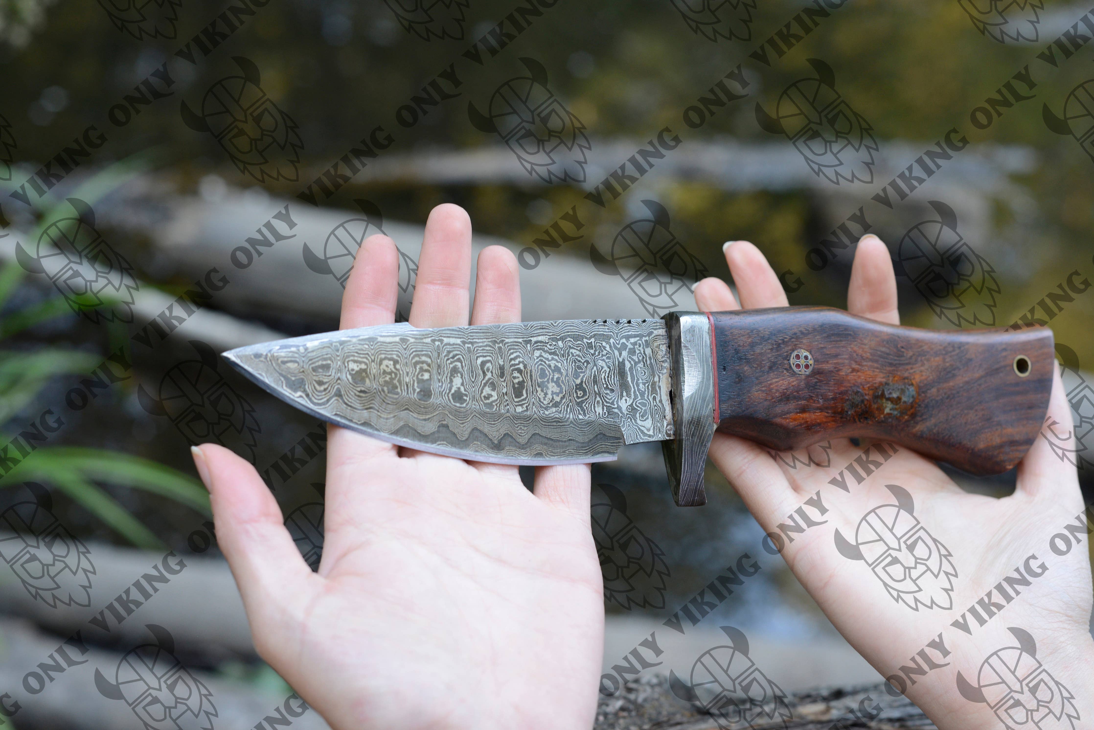 A person holds the Premium Quality Utility Knife: Damascus Knife, featuring a patterned blade and a wooden handle, set against a blurred natural backdrop with watermarks overlaying the image.