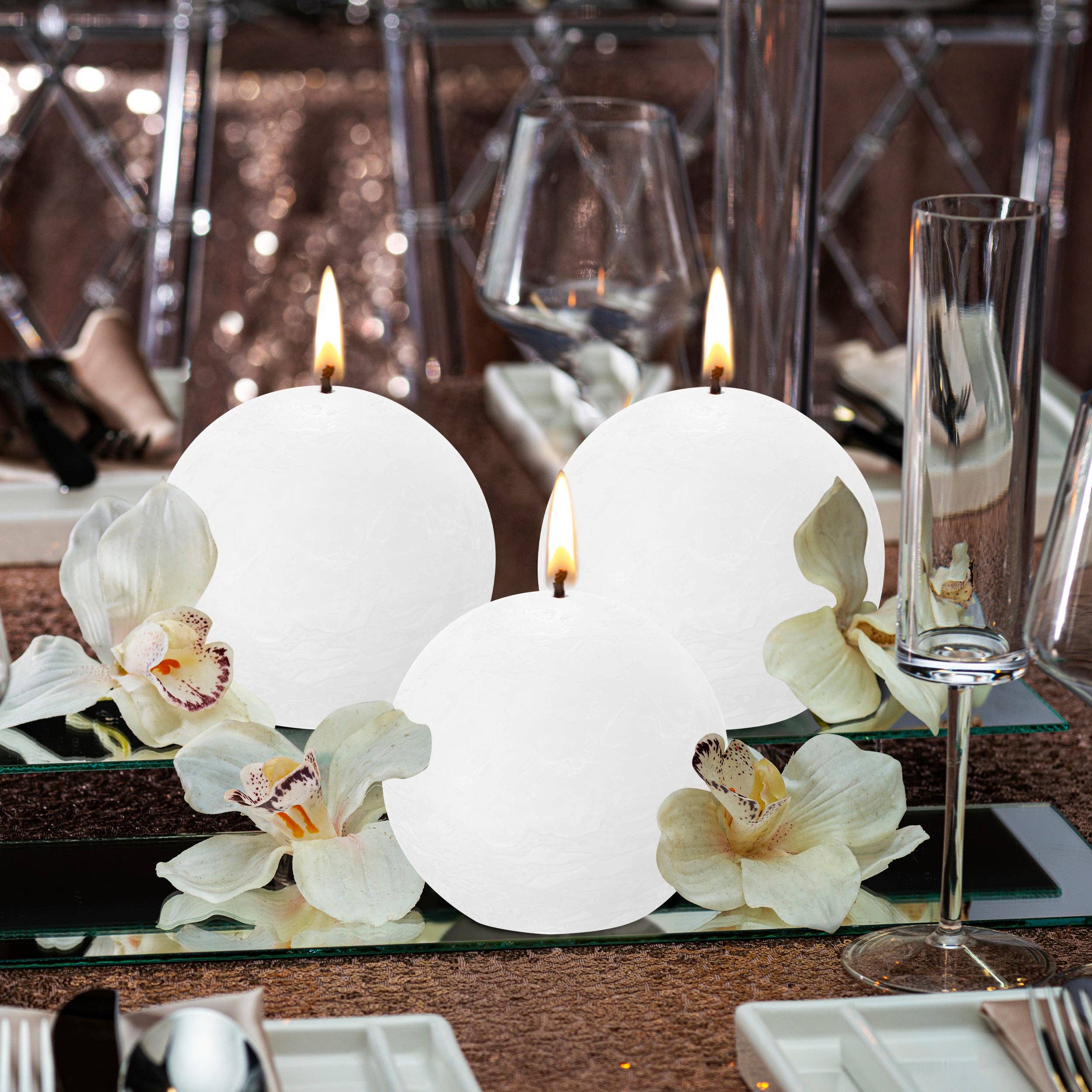 A trio of Cloudy White Rustic 3" Ball Candles, made from plant-based wax, are beautifully lit on a reflective surface and encircled by delicate white orchids. In the background, elegant crystal glassware and plates add a touch of sophistication.