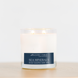 The Fjord & Fable Sea Minerals Candle, an 8oz bowl with 2 wicks and a 20+ hour burn time, fills the room with coastal scents of sea salt, eucalyptus, ozone, and moss. The candle sits on a light wooden surface.