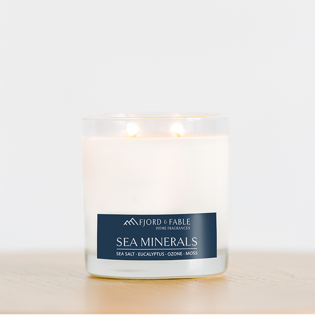 The Fjord & Fable Sea Minerals Candle, an 8oz bowl with 2 wicks and a 20+ hour burn time, fills the room with coastal scents of sea salt, eucalyptus, ozone, and moss. The candle sits on a light wooden surface.
