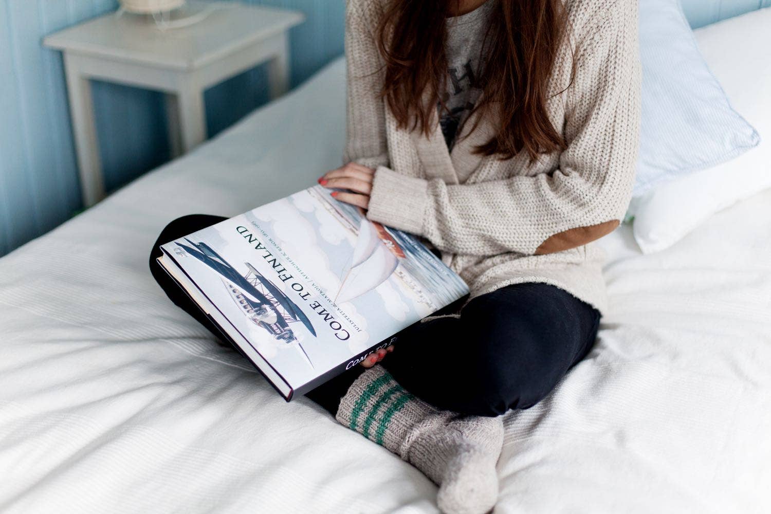 A person on a bed, in a sweater, black pants, and socks, holds the hardcover Book: Come to Finland, Coffee Table Book in English, featuring vintage travel posters.