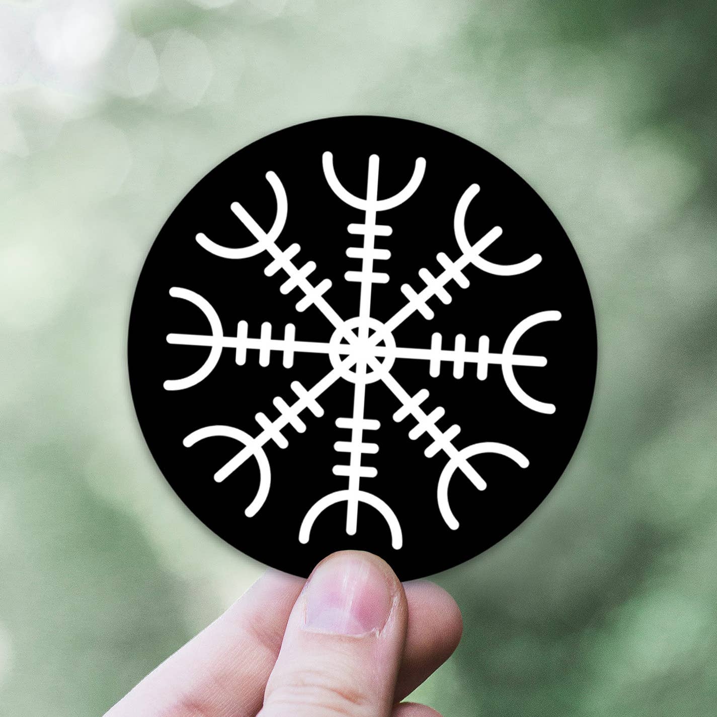 A hand holds a circular black sticker displaying a white Viking symbol known as the Helm of Awe, an ancient Norse protection symbol similar to the Vegvisir, against a blurred green background.