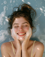 A young woman with closed eyes smiles while washing her face, holding the Eggwhite & Chamomile Facial Soap from the Gingham Gift Box (1.76 oz), lying back in water with bubbles.