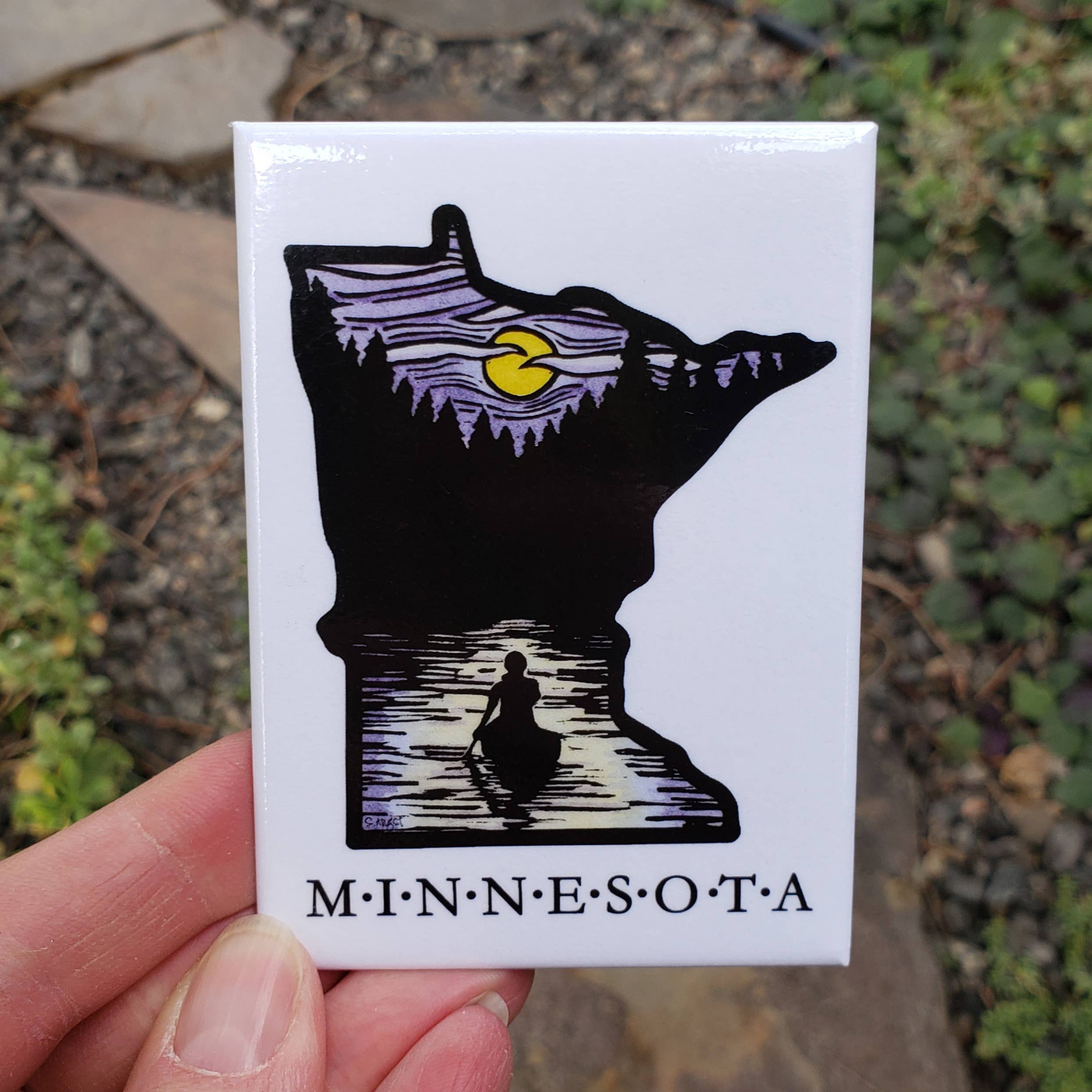 A hand displays the Minnesota Solitude Canoe Magnet, featuring a silhouette of a person paddling on a serene lake at sunset, framed by Minnesotas shape with the word Minnesota below.