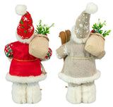 Two plush holiday figures in festive coats and hats, each holding a burlap sack and wooden skis, with greenery and red berries at their feet.