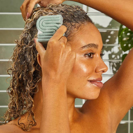 Standing in the shower, a person lathers their curls with Kitsh’s Rosemary & Biotin Volumizing Shampoo & Conditioner Bar Combo—enjoying sulfate-free care in convenient 2.7oz bars.