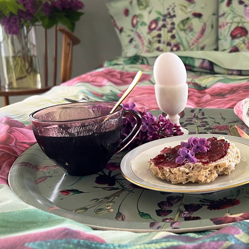 A round serving tray holding a cup of dark tea, a soft-boiled egg in an egg cup, and toast with jam, set against floral bedding in the background.