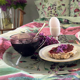 A round serving tray holding a cup of dark tea, a soft-boiled egg in an egg cup, and toast with jam, set against floral bedding in the background.