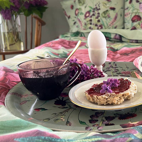 A round serving tray holding a cup of dark tea, a soft-boiled egg in an egg cup, and toast with jam, set against floral bedding in the background.