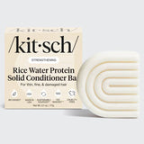 A box labeled "KITSCH Conditioner: Rice Water Protein Solid Conditioner Bar for Hair Growth 2.7oz" sits next to a white, curved, textured conditioner bar. The packaging highlights benefits for hair growth and added volume in thin, fine, and damaged hair—powered by Rice Tein NPNF—and displays icons indicating the product is bio-based, cruelty-free, and made in the USA.