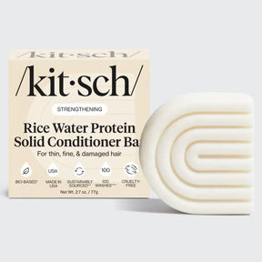 A box labeled "KITSCH Conditioner: Rice Water Protein Solid Conditioner Bar for Hair Growth 2.7oz" sits next to a white, curved, textured conditioner bar. The packaging highlights benefits for hair growth and added volume in thin, fine, and damaged hair—powered by Rice Tein NPNF—and displays icons indicating the product is bio-based, cruelty-free, and made in the USA.