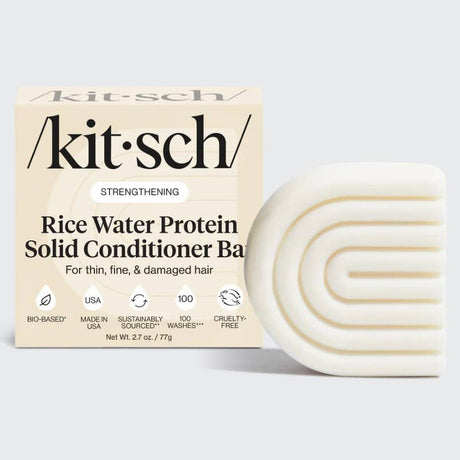 A box labeled "KITSCH Conditioner: Rice Water Protein Solid Conditioner Bar for Hair Growth 2.7oz" sits next to a white, curved, textured conditioner bar. The packaging highlights benefits for hair growth and added volume in thin, fine, and damaged hair—powered by Rice Tein NPNF—and displays icons indicating the product is bio-based, cruelty-free, and made in the USA.