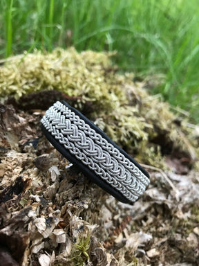 The Traditional Sami Bracelet #5B features black reindeer leather and pewter-silver thread, displayed on a mossy log with green grass in the background. Available in 3 sizes.