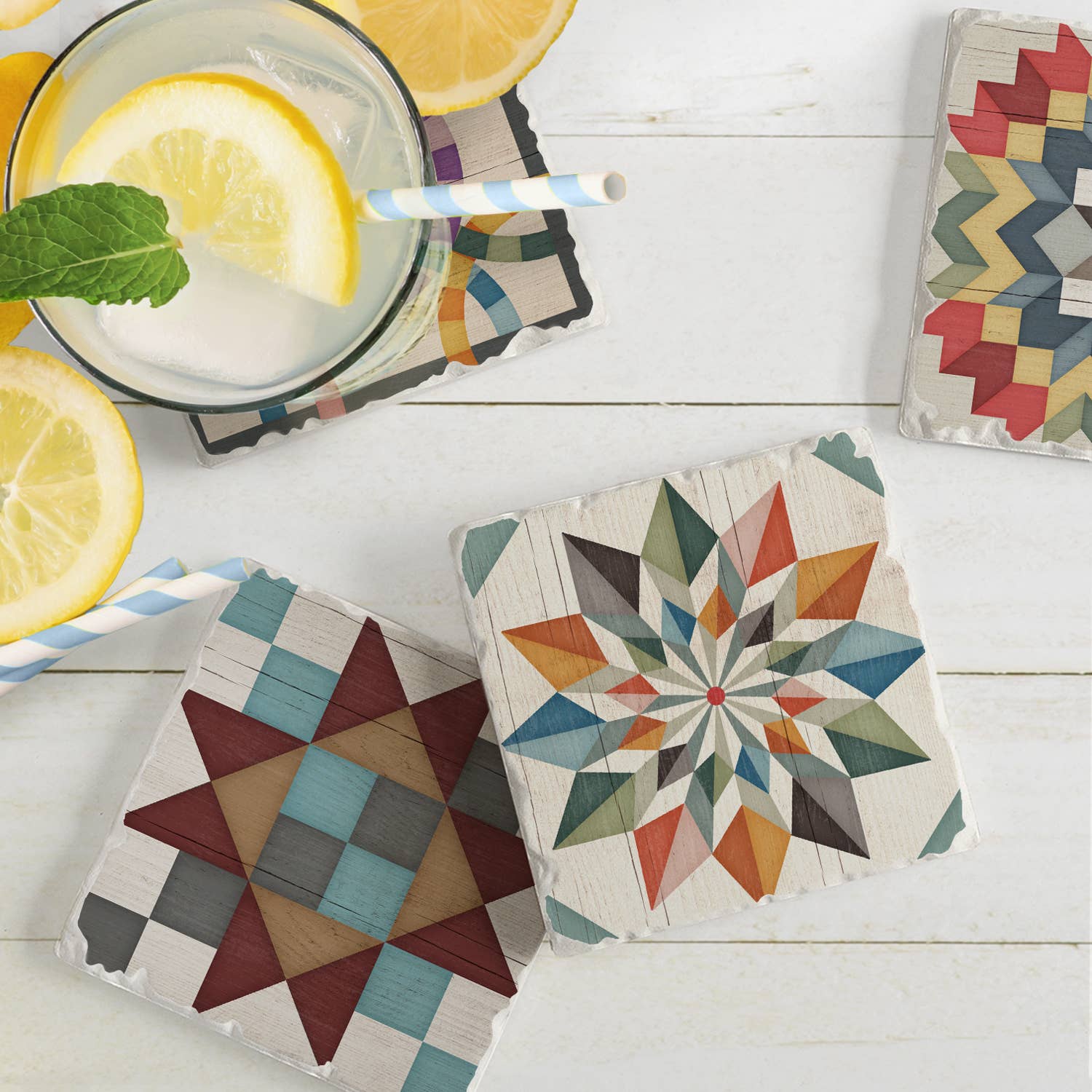 A glass of lemonade with a mint leaf and striped straw sits on a vibrant Barn Quilt Signs Stone Coaster with cork backing. Nearby are three more coasters featuring geometric designs, all resting on a white wooden surface.