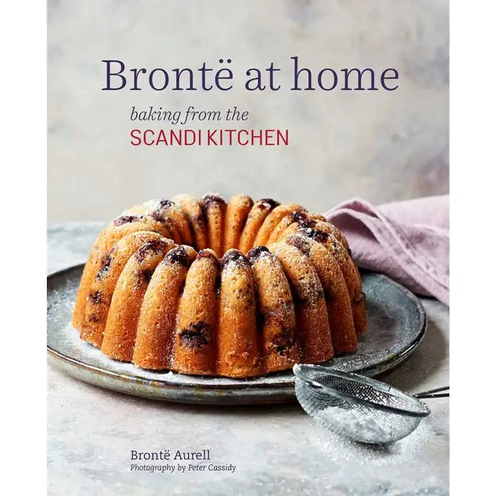 Cover of Bronte at Home - Baking from the Scandikitchen by Bronte Aurell, with a sugar-dusted Bundt cake, a sieve, and a pink cloth.