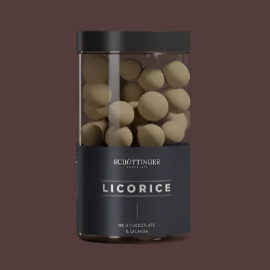 A clear jar labeled “Schottinger Chocolate Licorice Milk Chocolate & Salmiak 250g 8.8 oz” holds round, pale-coated candies with a black lid, set against a plain brown background.