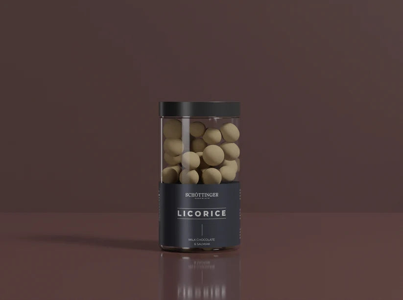 A clear jar labeled “Schottinger Chocolate Licorice Milk Chocolate & Salmiak 250g 8.8 oz” holds round, pale-coated candies with a black lid, set against a plain brown background.