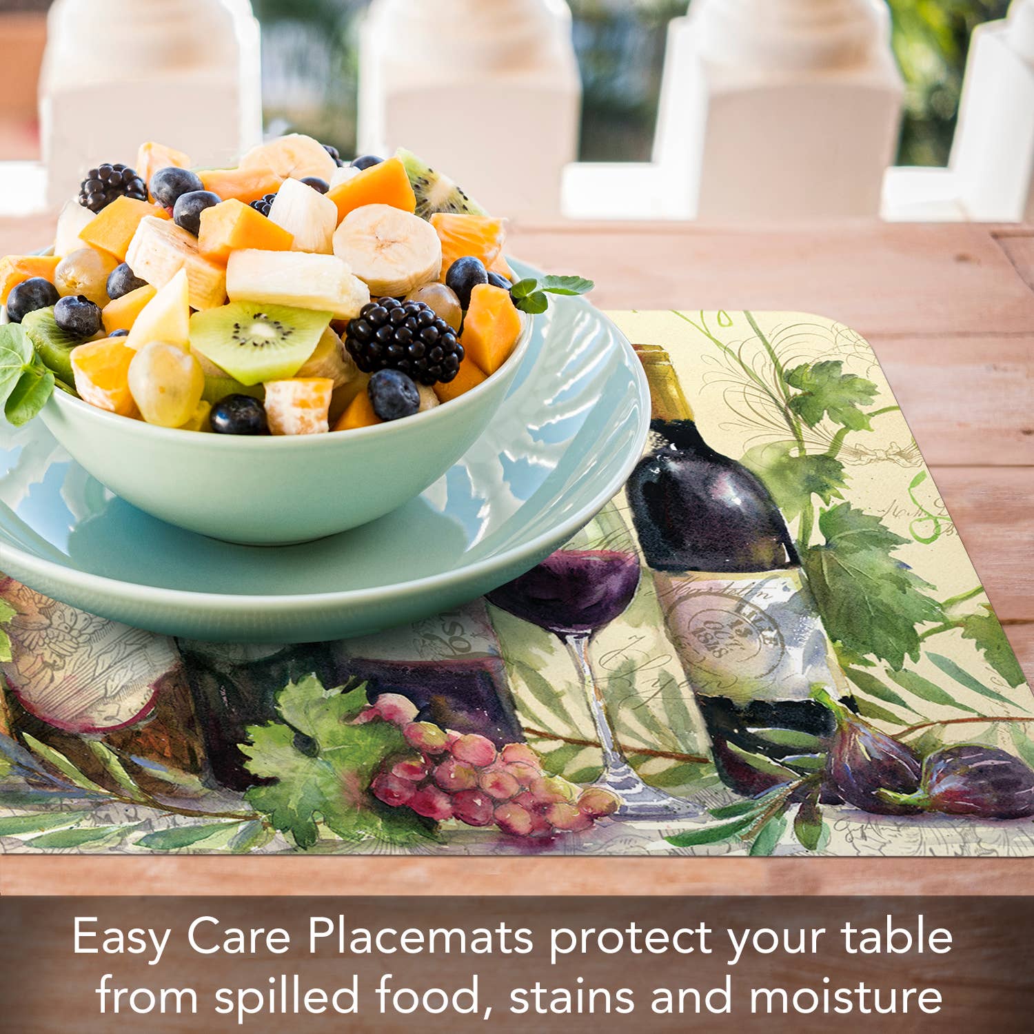 A bowl of assorted fruits rests on a decorative placemat featuring a rustic wine bottle design. Text reads: Rustic Wine Reversible Rectangular Plastic Placemat, easy care, BPA free, designed to protect your table from spills, stains, and moisture.