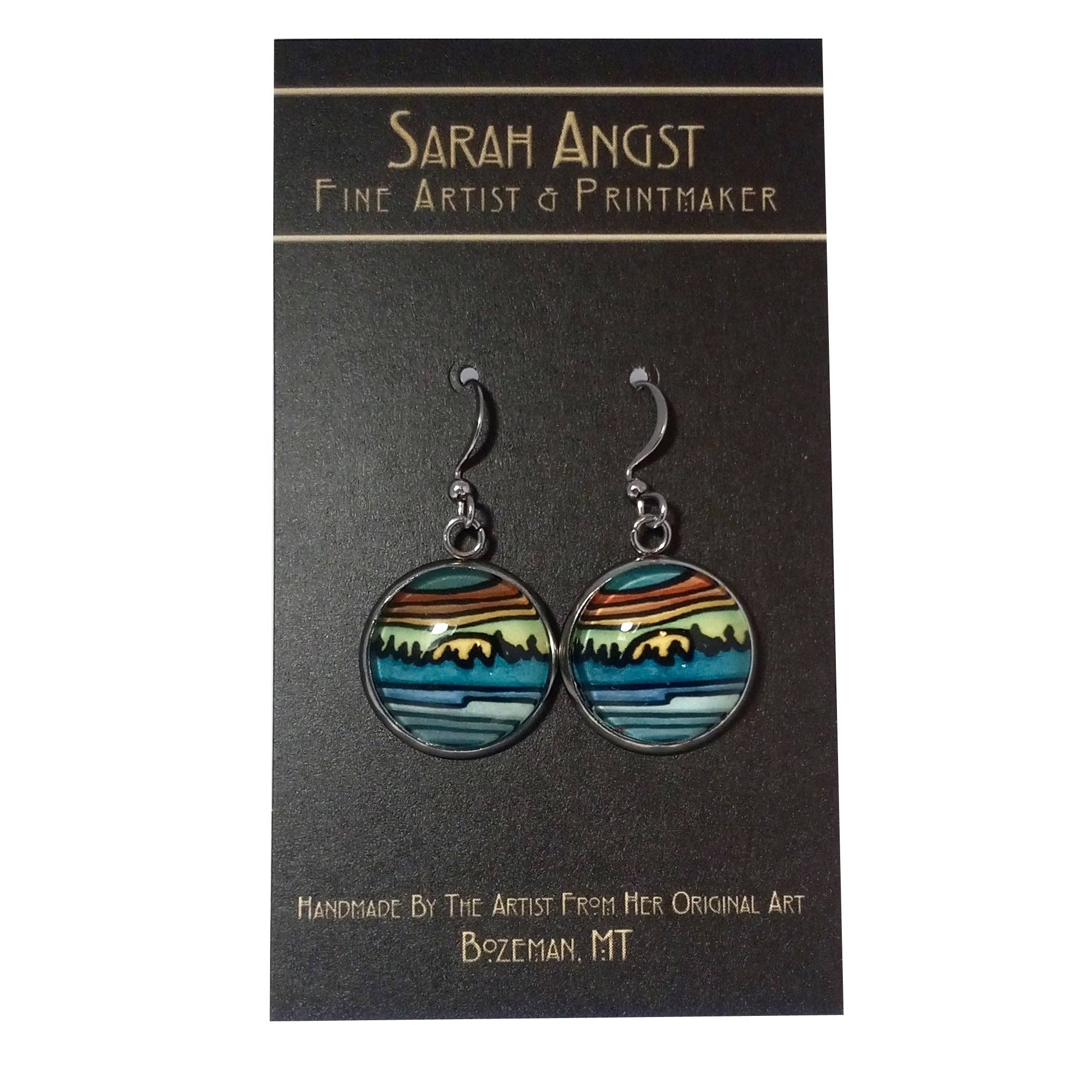 Earrings titled "Dock View by Sarah Angst" feature a blue, green, and orange landscape design and are displayed on a card labeled "Sarah Angst Art, Fine Artist & Printmaker, Bozeman, MT," reflecting the vibrant artistry of the region.