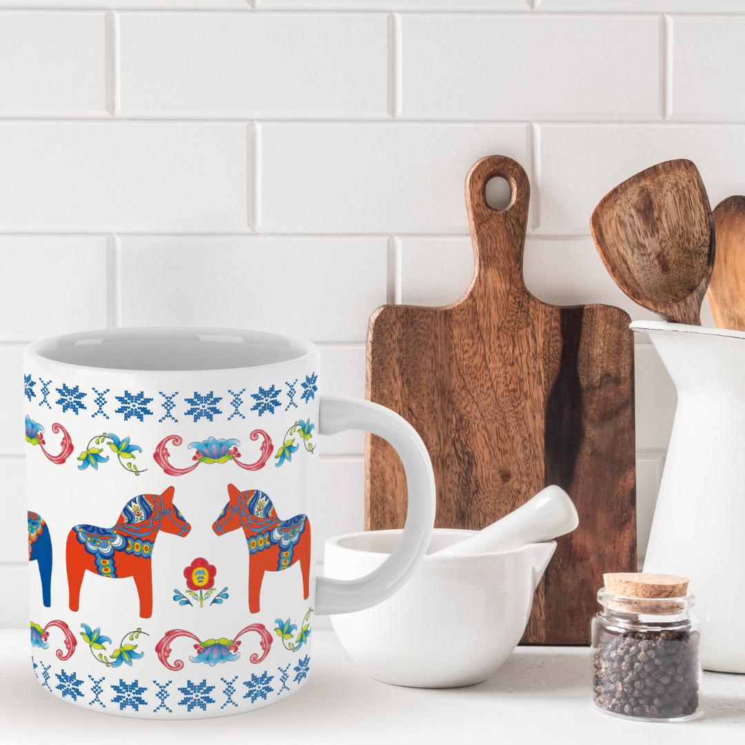 A white ceramic coffee cup featuring a vibrant Dala Horse Rosemaling design is placed on a kitchen counter, surrounded by cutting boards, a mortar and pestle, and spice jars, capturing the essence of Scandinavian charm.