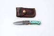 The Titan International Knife: Turquoise Damascus Folding Knife – Hand Engraved Bolster Edition features a turquoise inlay and Damascus blade, shown with a stitched brown leather sheath and brass snap, all on a white background.