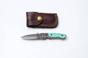 The Titan International Knife: Turquoise Damascus Folding Knife – Hand Engraved Bolster Edition features a turquoise inlay and Damascus blade, shown with a stitched brown leather sheath and brass snap, all on a white background.