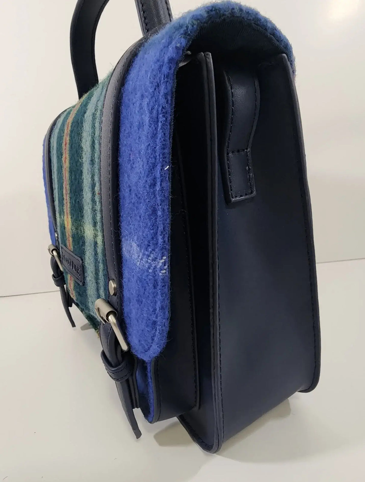 A side view of the Bag: Nova Scotia Patsy Handbag shows its structured shape, blue and green plaid fabric, blue wool trim, leather details, and top handle.