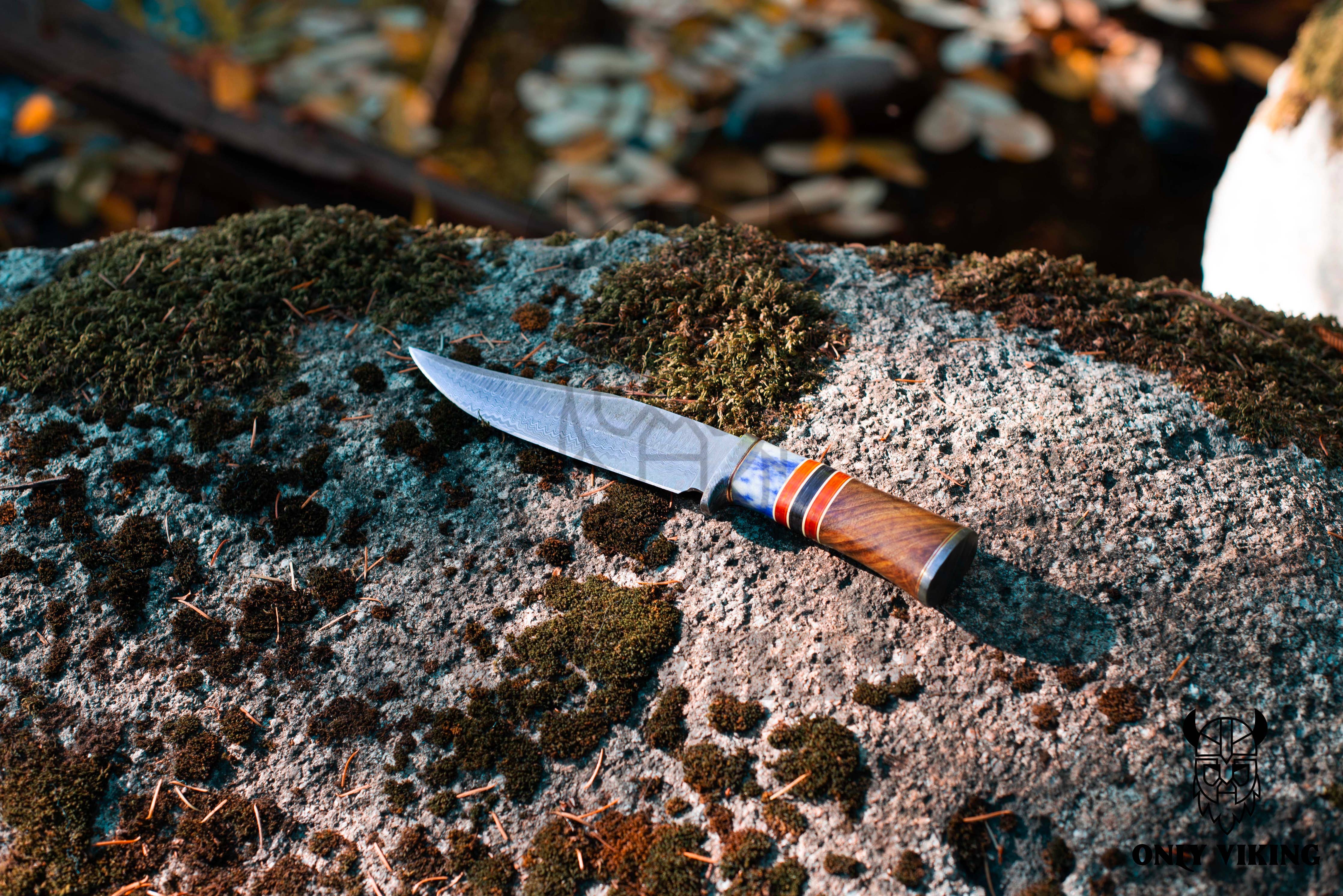 A Damascus Steel Hunting Knife with a wooden handle and intricately patterned blade rests on a moss-covered rock outdoors.