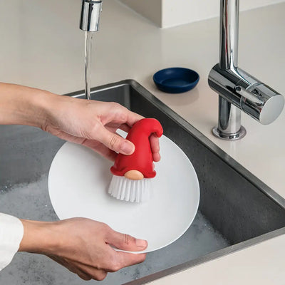 Someone washes a white plate under running water using the OTOTO Brush: Beardy Tomte — a 4" tall, gnome-shaped dish scrub brush with a red hat and 3.25" diameter dish, which is dishwasher safe.