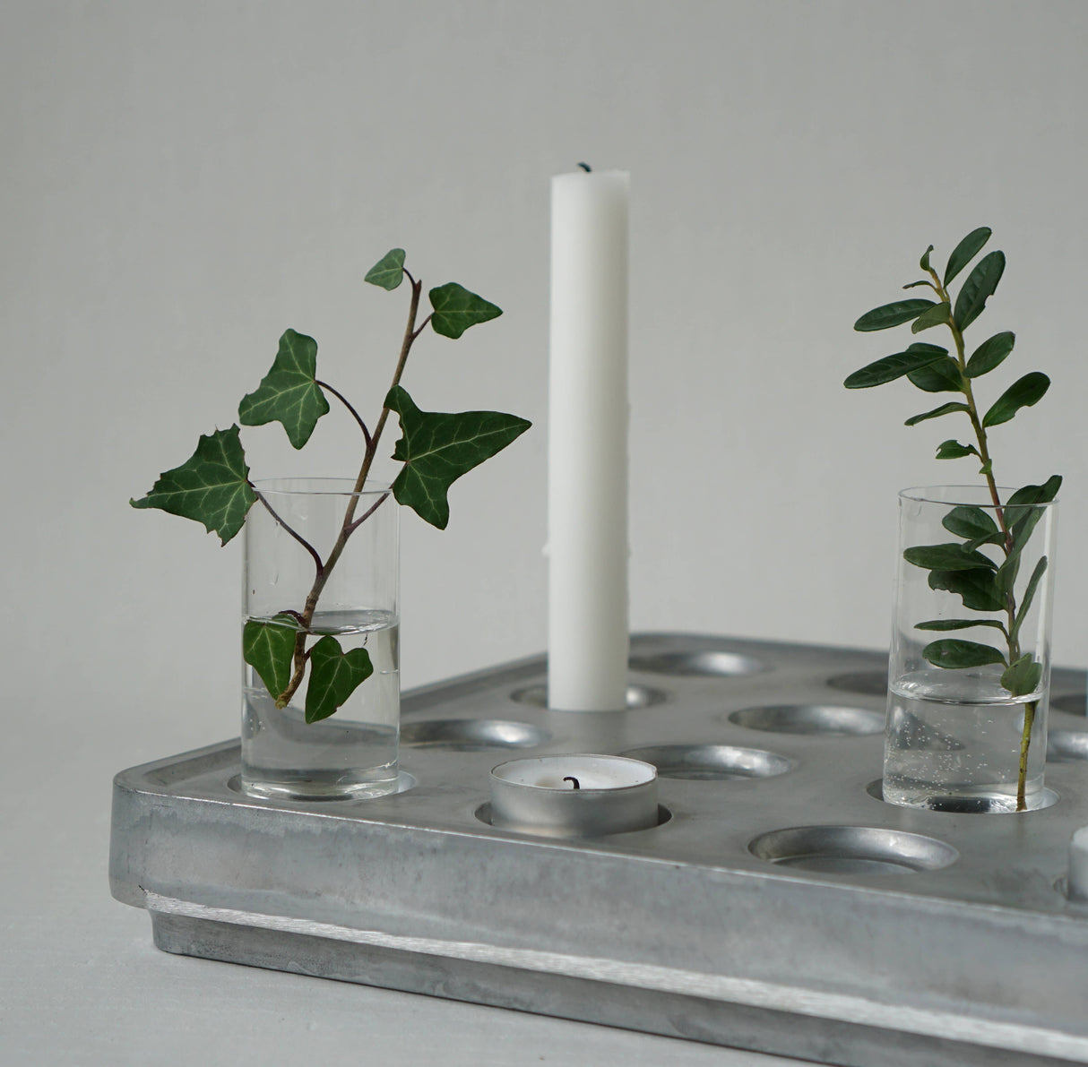 The Candle Holder: Mini Vases for Stumpastaken Candleholder 2-pack, with green leafy stems, pairs with a white pillar candle and tealight on a metal tray—an ideal table decoration against a light gray background.