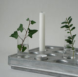 The Candle Holder: Mini Vases for Stumpastaken Candleholder 2-pack, with green leafy stems, pairs with a white pillar candle and tealight on a metal tray—an ideal table decoration against a light gray background.