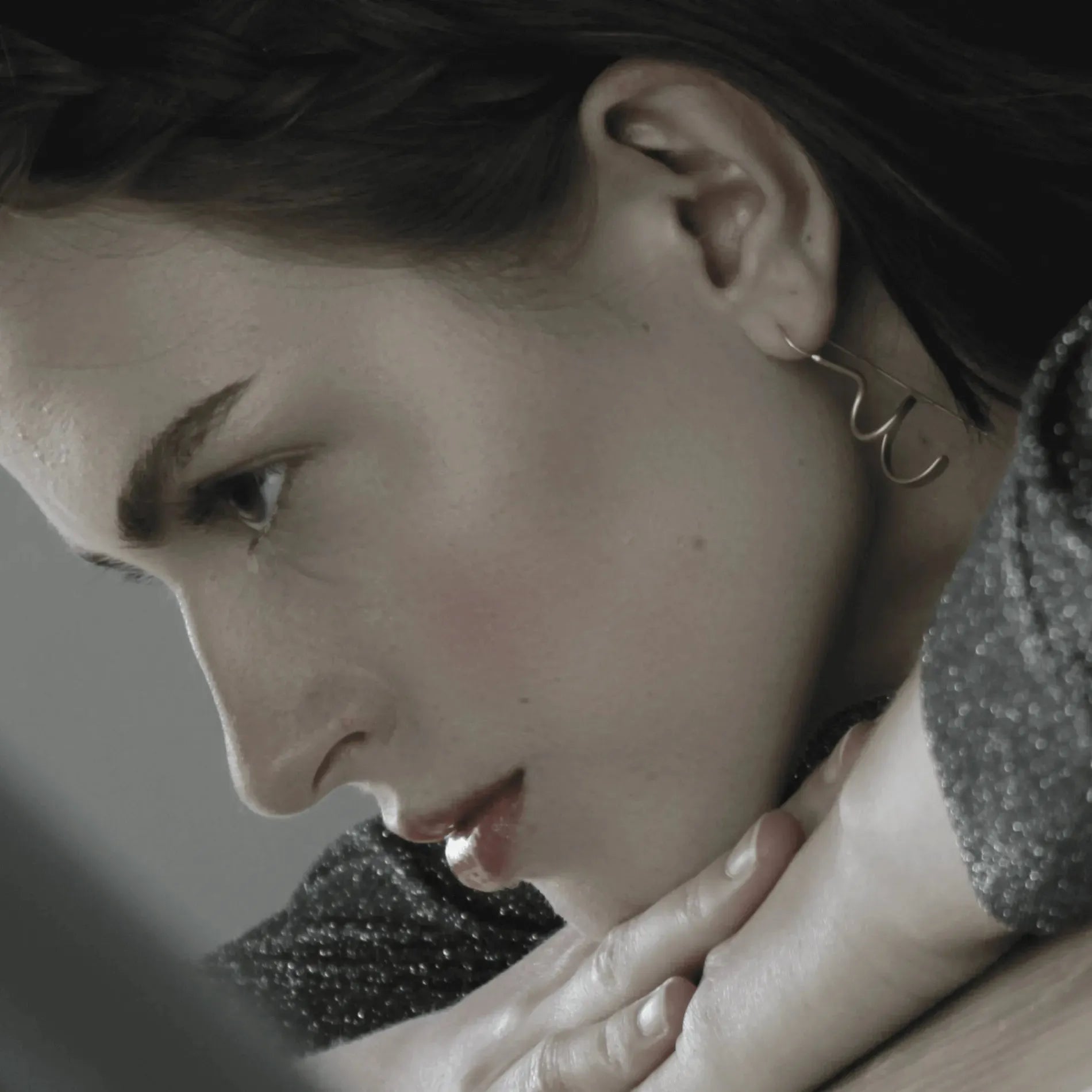 A person with light skin and dark hair rests their chin on crossed hands, gazing downward; they are wearing Dansk Copenhagen Tara Mini Spinning Silver Earrings and a textured top.