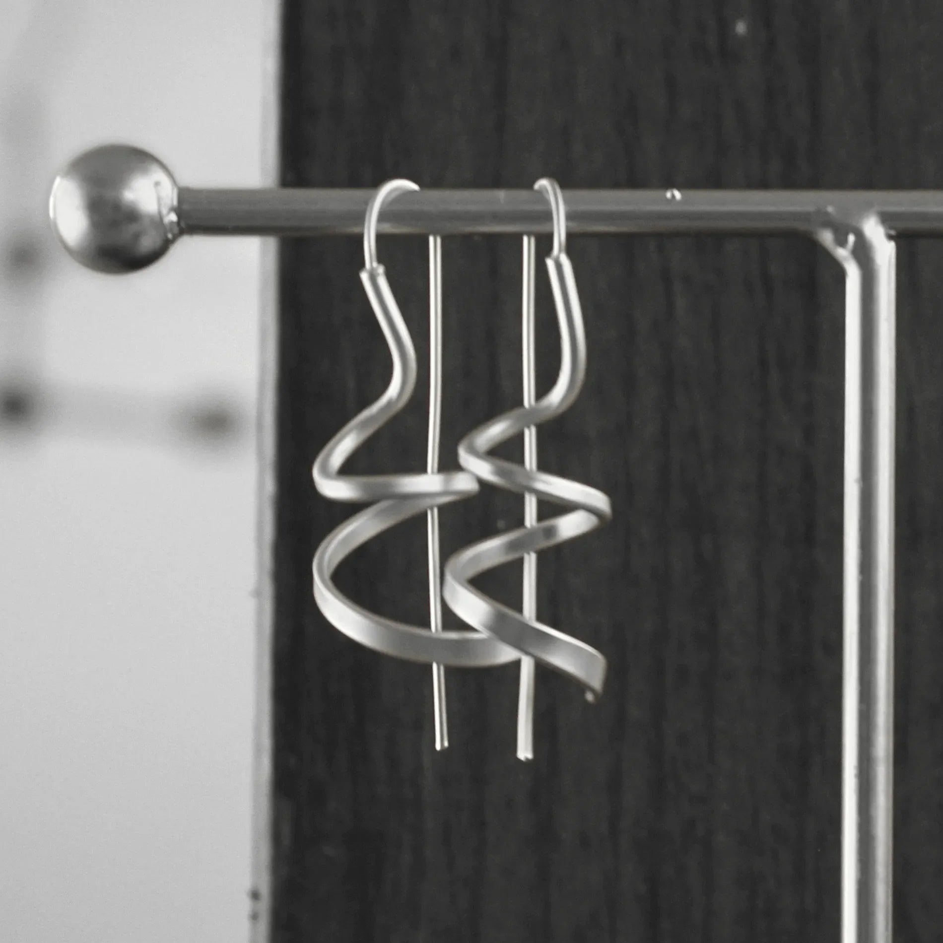 A pair of Dansk Copenhagen Tara Mini Spinning Silver Earrings hang on a metal display stand against a dark wooden background.