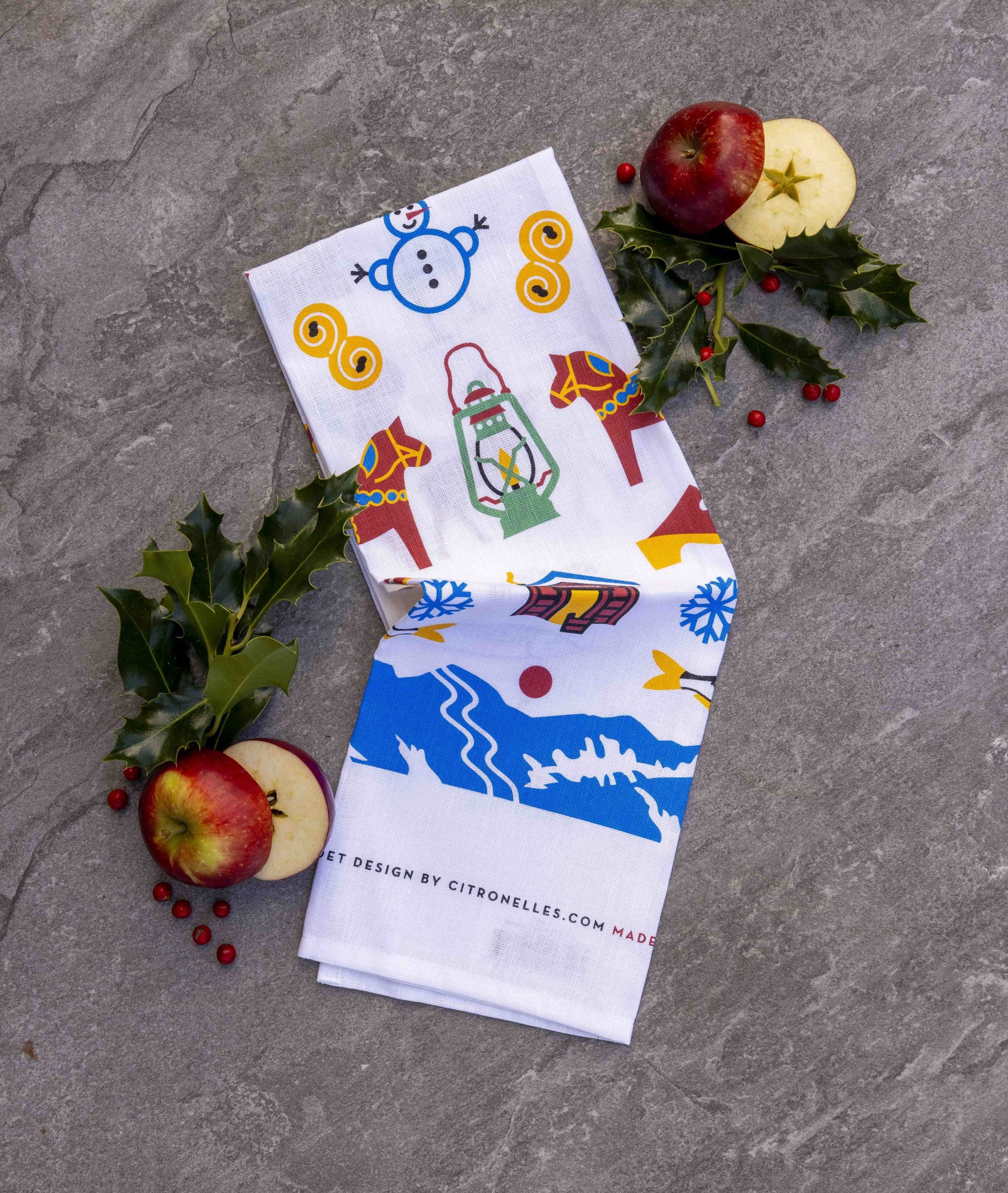 The Tea Towel: Swedish Winter Traditions – Cotton/Linen Blend (50 × 70 cm) with festive holiday patterns is displayed on a gray surface, surrounded by apples, holly leaves, and red berries.