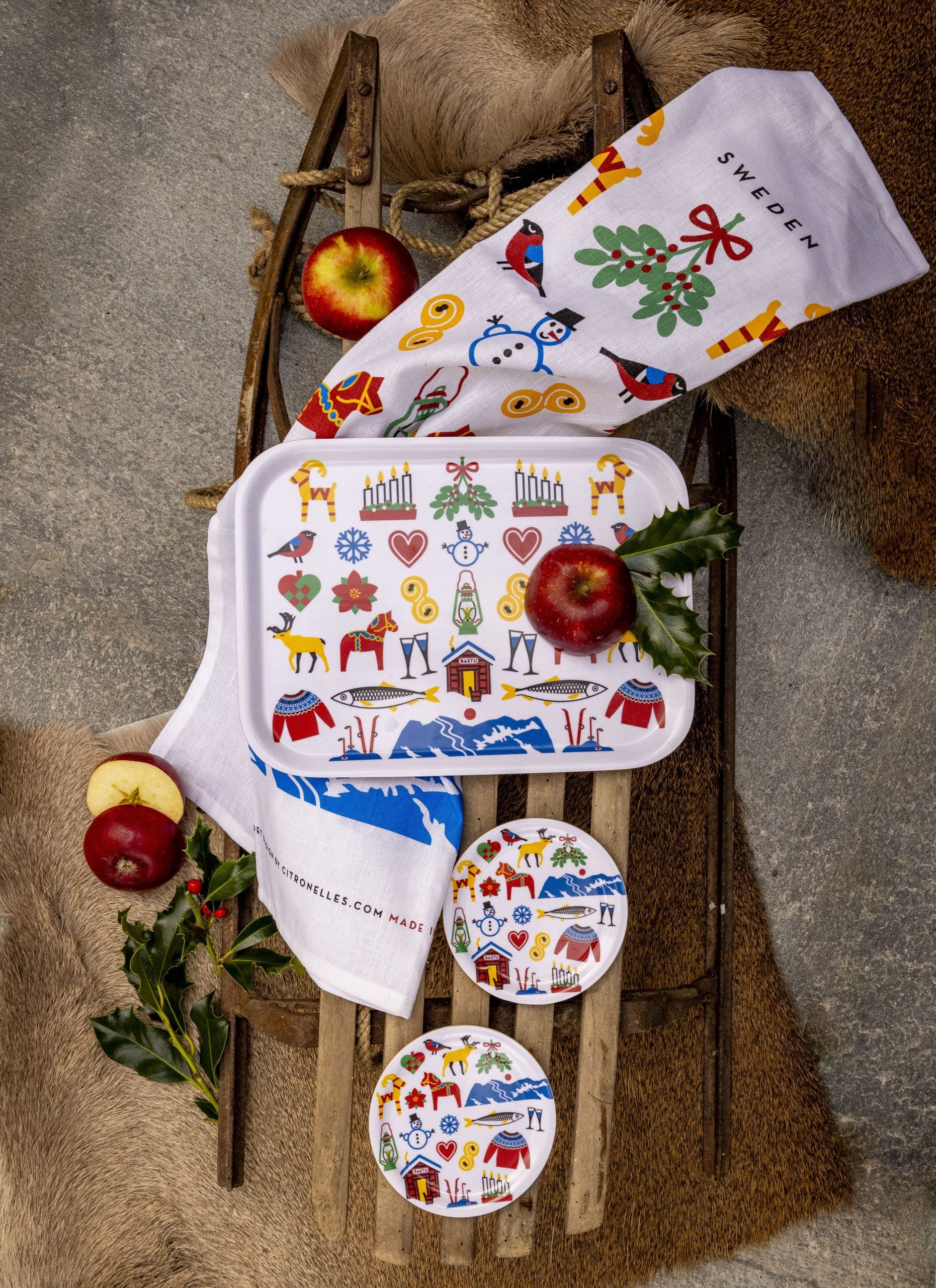 A tray, coasters, and the Tea Towel: Swedish Winter Traditions (cotton/linen blend, 50×70 cm) with colorful Swedish folk art designs are displayed on a fur-covered sled, accented by apples and greenery.