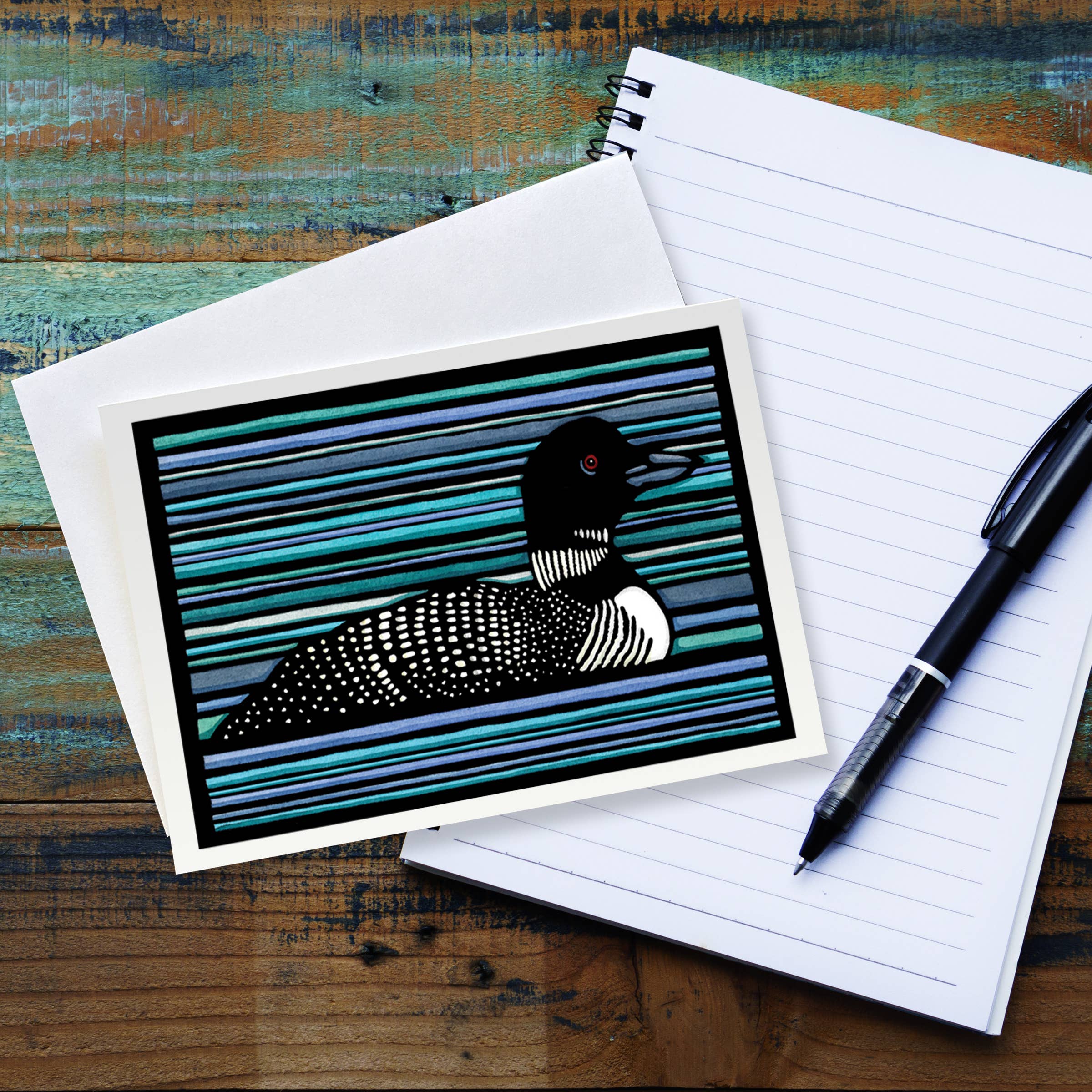 The Card: Loon 5x7 Greeting Card with Envelope Blank Inside rests on a lined notepad with a pen atop a rustic wooden surface. This card features a charming loon illustration, perfect for any occasion.